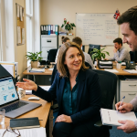 HR director reviewing an AI dashboard on a laptop in a small UK office, mid-conversation with a colleague.